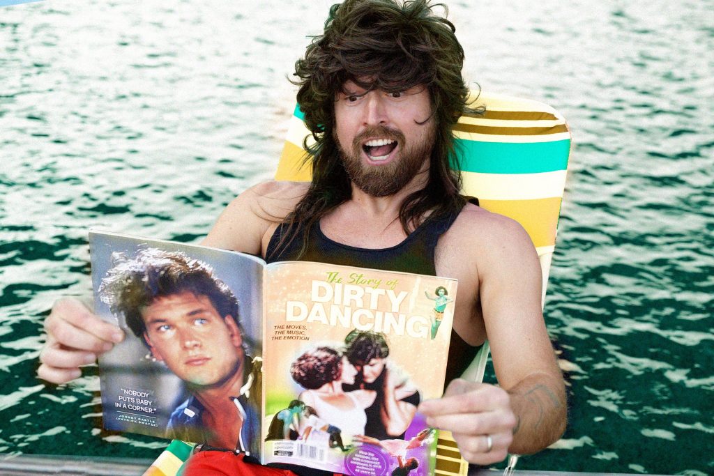 Promotional photo for "Swayze" which sees Iglu & Hartly frontman, Jarvis Anderson, in a wig, lying on a deck chair, reading a Dirty Dancing magazine.