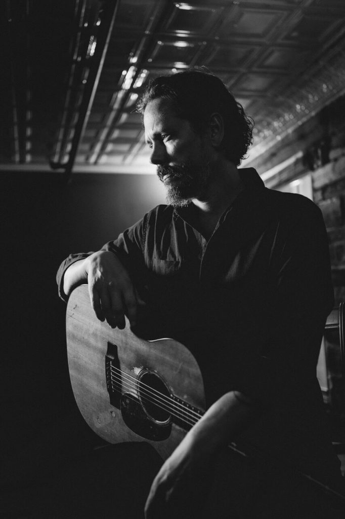 Promotional photo for "Big Love" with Wildo DM, which is a black and white photo of John Paul White looking to the left, sitting down with his guitar sat on his lap and one arm holding the body of the guitar.