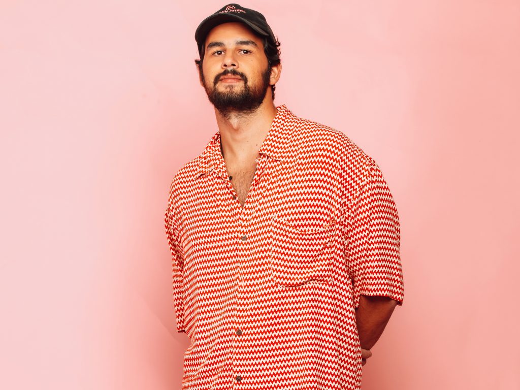 Promotional photo for "Sky Father" in collaboration with Antdot and Beacon Bloom, which sees Maz (BR) standing in front of a baby pink backboard, wearing a salmon-white checkered shirt and a flat cap.