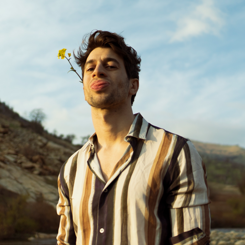 Promotional photo for "Unconditional" which sees Mike Ruby sticking his tongue out, with a daisy in his hair, and he is wearing a vertical-striped white and brown shirt. The countryside hills can be seen in the background.