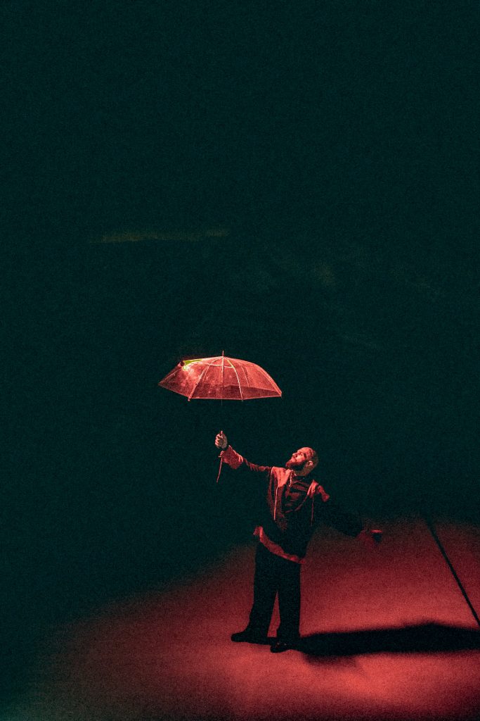 Promotional image for "Reckless" which sees a long shot of Moh Flow lit in a red spotlight, holding out an umbrella.