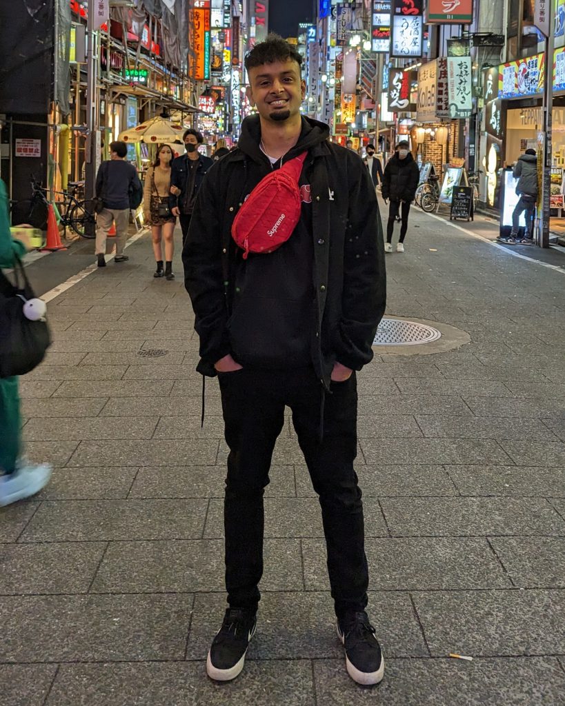 Promotional photo for "Ocean Drive" which sees Poet Initiative standing on a pedestrian shopping street with the shop lights illuminating the street. He is wearing all black, has his hands in the pockets of his black skinny jeans, and he has a red cross-body bag over his black hoodie.