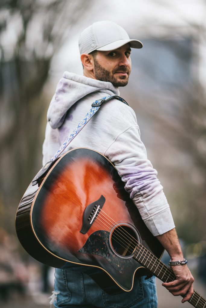 Promotional photo for "Awol" which sees Rob Roth wearing blue jeans and a grey hoodie with a matching snapback cap, looking over his shoulder at the camera with his guitar strap slung over one shoulder with the guitar held against him.