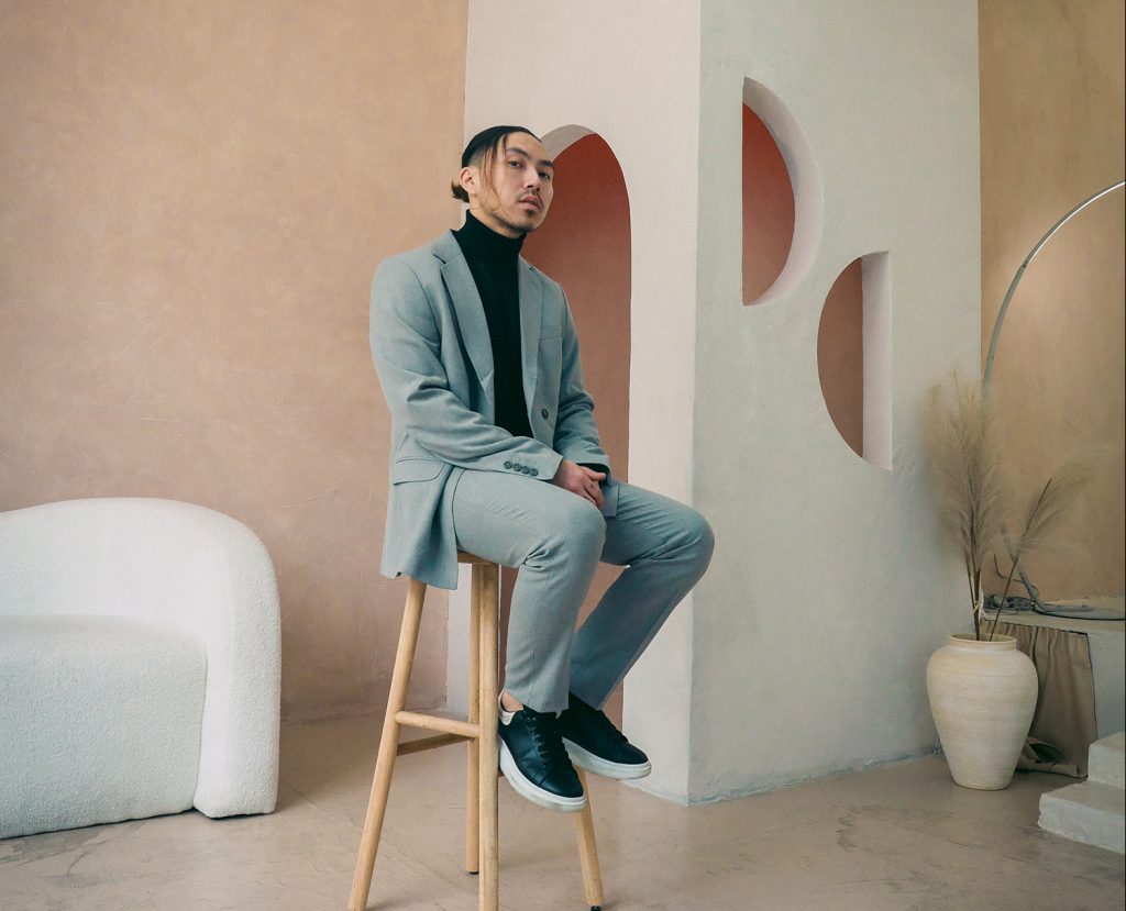 Promotional photo for "broken: playlist II" which sees VeeAlwaysHere sitting on a wooden stool wearing a grey two-piece suit with a black jumper and black trainers, while his hair is tied back in a bun. He's sitting in a brightly-lit beige-pink room with an interesting backroom square with white walls.