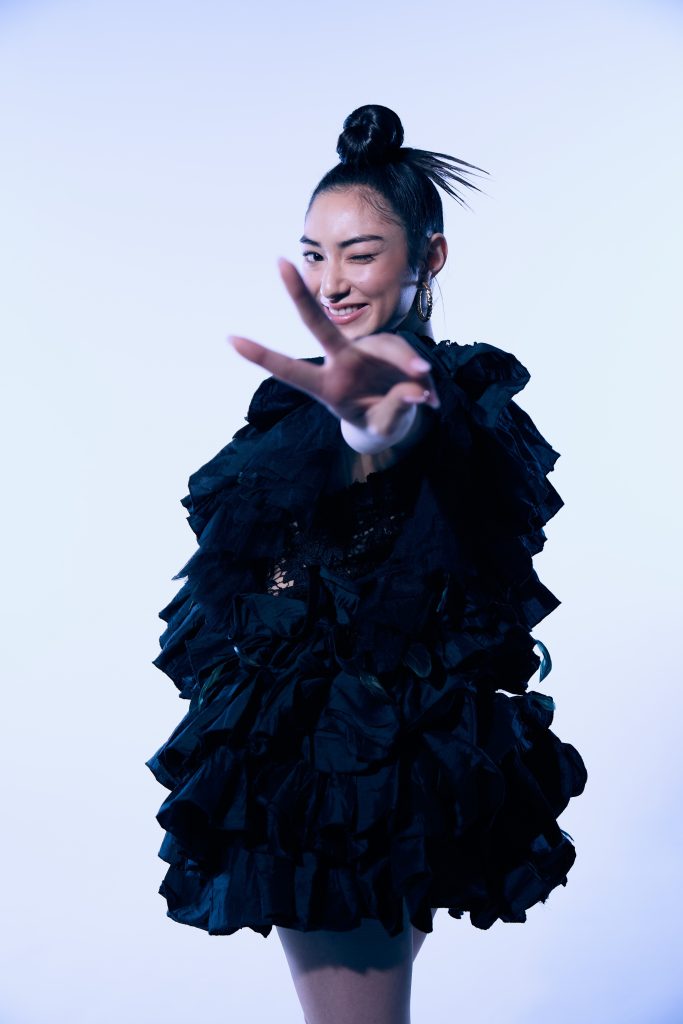 Professional photo image of Anna Aya posing wearing a ruffled oversized black dress, with her hair in a bun on top of her head. She's facing to the left with her head turned towards the camera and she's got her hand extended and throwing the peace sign.