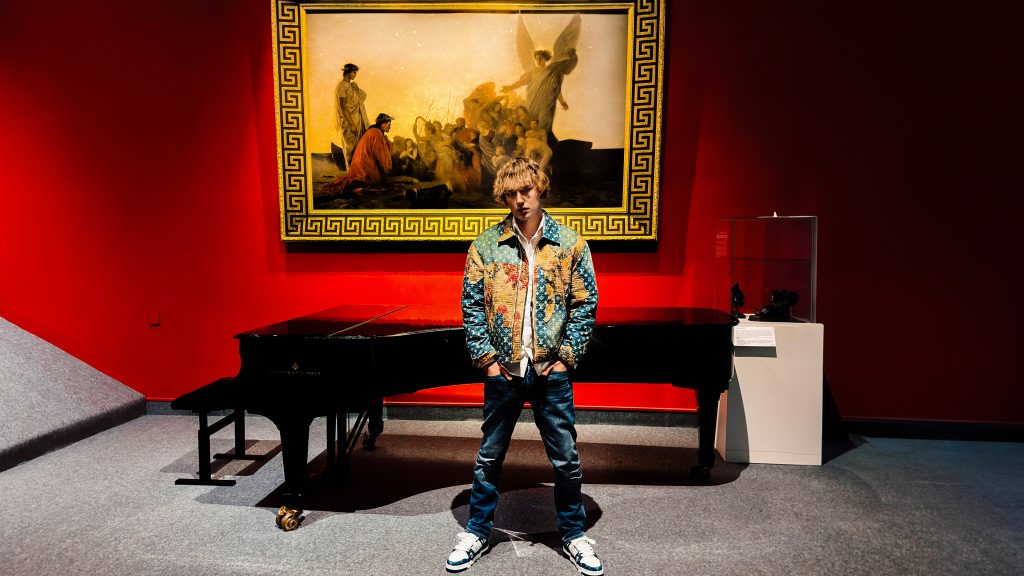 Promotional image for "HISTORY" which sees Alex Shade standing in front of a black piano, with an art painting behind him that's hanging on royal red walls.